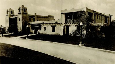 Historic photo of the New Mexico State Building featuring adobe-style architecture, flat roofs, and rounded corners, with a sidewalk, streetlamp, and a person walking nearby. [gen-ai]