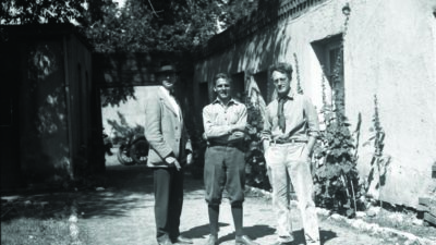 Three men stand in a sunlit outdoor corridor next to a stucco building, with plants growing along the wall and a motorcycle visible in the background. [gen-ai]