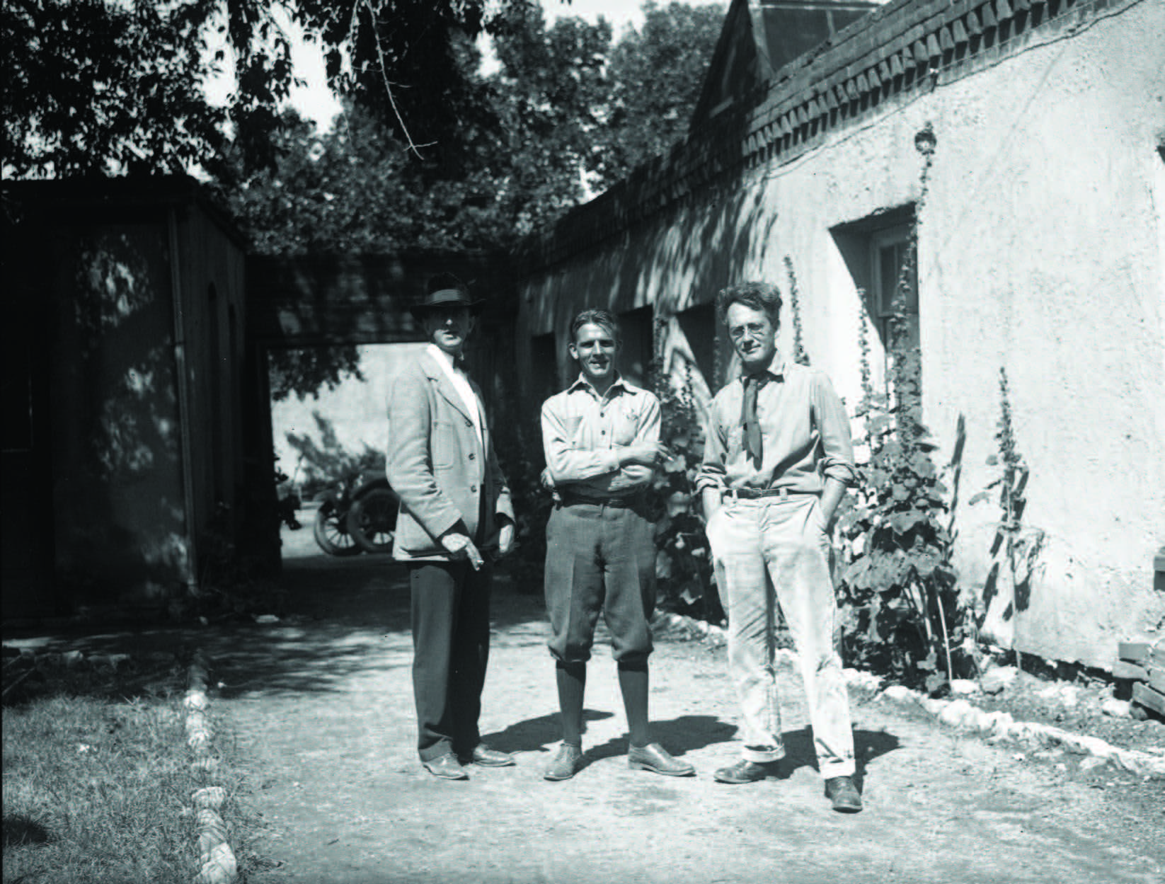 Three men stand in a sunlit outdoor corridor next to a stucco building, with plants growing along the wall and a motorcycle visible in the background. [gen-ai]