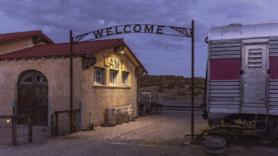 A vintage train station with a “WELCOME” sign, an old building labeled “LAMY,” and part of a silver train car visible at dusk. [gen-ai]
