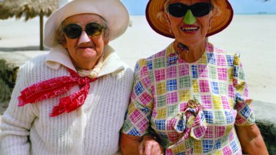 Two elderly women in sun hats and sunglasses stand arm in arm on a beach with straw umbrellas visible in the background. [gen-ai]