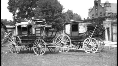 Two old wooden stagecoaches with large wheels are parked on grass; trees and a building with arched windows appear in the background. [gen-ai]