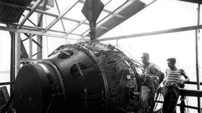 Two men stand next to a large, spherical device covered with wires inside a partially constructed building with exposed beams. [gen-ai]