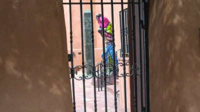 Wrought iron gate in an adobe wall opens to a courtyard, with a person in colorful clothing walking past in the background. [gen-ai]