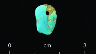 A turquoise stone pendant with a small drilled hole, placed against a black background with a centimeter scale below showing 0 to 3 cm. [gen-ai]