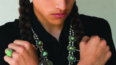 Person with long braids wears a black shirt, silver and turquoise jewelry, and crosses their arms over their chest while looking downward. [gen-ai]