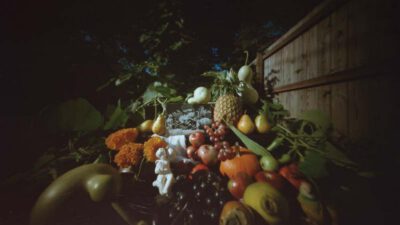 A variety of fruits, vegetables, flowers, and small figurines arranged outdoors on a table next to a wooden fence with greenery in the background. [gen-ai]