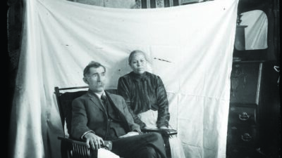 A man and woman pose for a portrait in front of a white sheet in a decorated room, with a dog lying on the floor in the foreground. [gen-ai]