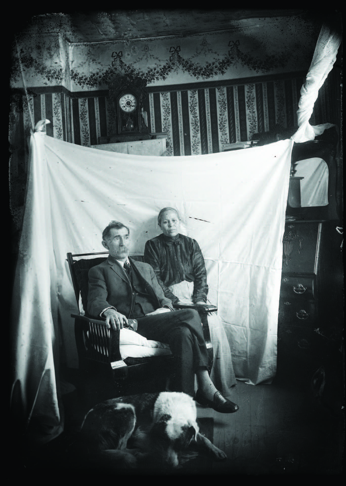 A man and woman pose for a portrait in front of a white sheet in a decorated room, with a dog lying on the floor in the foreground. [gen-ai]