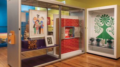 A museum display case with colorful graphic art, a framed photograph, a red brick panel, and decorative objects on wooden flooring. [gen-ai]