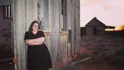 A person in a black dress stands with arms crossed against a weathered wooden building in a rural, grassy area at sunset. Another building is visible in the background. [gen-ai]