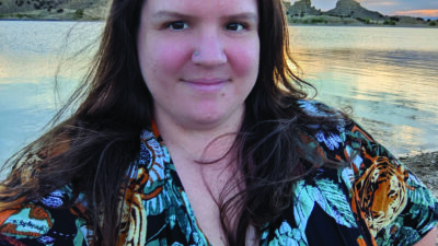 A woman with long brown hair stands by a lake at sunset, wearing a floral-patterned dress. The sky is partly cloudy with hills visible in the background. [gen-ai]
