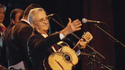 An older man in a suit holds a small guitar and gestures toward a microphone on stage, with other musicians in the background. [gen-ai]
