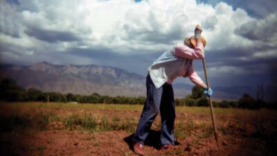 A person leans on a hoe in a field under a cloudy sky; text reads, “If There’s Squash Bugs in Heaven, I Ain’t Staying” by Stacia Spragg-Braude. [gen-ai]