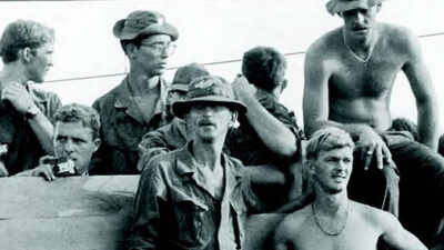 A group of soldiers in military attire and hats, some shirtless, sitting and standing together outdoors, likely during wartime. [gen-ai]