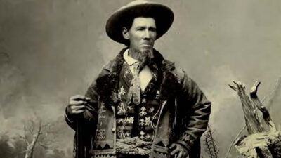 A man in old Western attire stands outdoors, wearing a wide-brimmed hat, patterned vest, and coat, with one hand resting on his hip and the other holding an object. [gen-ai]