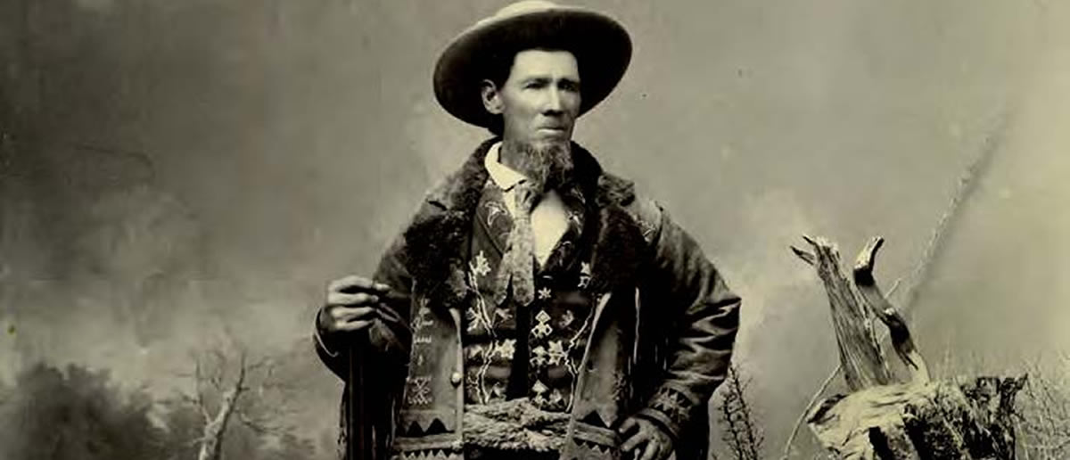 A man in old Western attire stands outdoors, wearing a wide-brimmed hat, patterned vest, and coat, with one hand resting on his hip and the other holding an object. [gen-ai]
