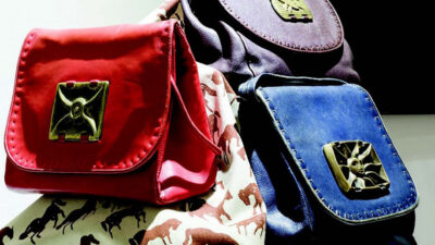 Three small handbags in red, purple, and blue with metallic clasps are displayed on a fabric printed with horse silhouettes. [gen-ai]