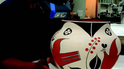 A person wearing gloves and a mask examines a painted ceramic pot with red, black, and white geometric and bird designs on a table. [gen-ai]