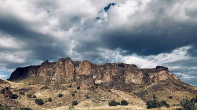 A rocky mountain range rises above dry grassland and scattered shrubs under a partly cloudy sky. [gen-ai]