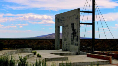A concrete outdoor stage with a large, rectangular wall structure and steel cables stands against a desert landscape and distant mountains under a partly cloudy sky. [gen-ai]