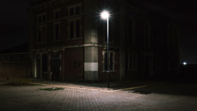 An old, dark building stands on a corner at night, illuminated by a single streetlamp on an empty, cobblestone street. [gen-ai]