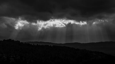 Sun rays break through dark clouds, illuminating patches of a forested landscape with rolling hills in a black and white scene. [gen-ai]