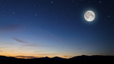 A full moon shines in a starry night sky above silhouetted mountains with a faint orange glow on the horizon. [gen-ai]