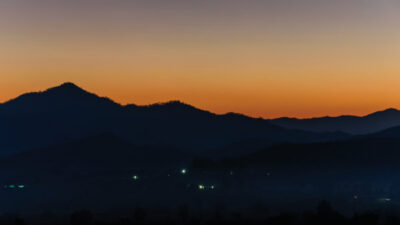 Silhouetted mountains at dusk with a gradient orange to blue sky and small lights scattered in the valley below. [gen-ai]