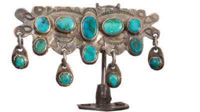 A silver decorative piece with engraved patterns, set with seven turquoise stones and small turquoise dangles, displayed on a stand against a white background. [gen-ai]