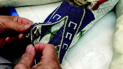 A person’s hands sew or repair a blue, white, and green beaded textile item on a padded white surface. [gen-ai]