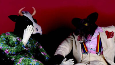 Two people wearing animal costumes, one as a cow in a green dress and the other as a wolf in a suit with rainbow suspenders, sit on a couch against a red background. [gen-ai]