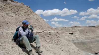 Person wearing outdoor gear and a backpack sits on a rocky slope, examining an object in their hands, under a partly cloudy sky in a barren landscape. [gen-ai]