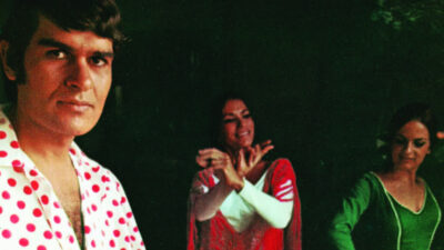 A man in a white shirt with red polka dots stands in the foreground, while two women in colorful outfits dance in the background. [gen-ai]