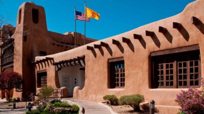 Adobe-style building with wooden accents, two flags (USA and New Mexico) on the roof, blue sky, and landscaping in front. [gen-ai]