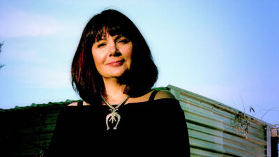 A woman with shoulder-length dark hair, wearing a black top and a large necklace, stands outdoors in front of a corrugated metal structure. [gen-ai]