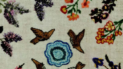 Embroidered fabric featuring colorful flowers, birds in flight, and a central circular blue pattern on a beige background. [gen-ai]