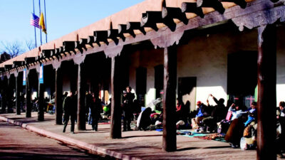 People sit and display goods for sale under the portico of a historic adobe building with wooden beams, with flags flying above. [gen-ai]