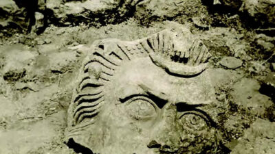 A large stone sculpture of a face partially buried in the ground among rocks and dirt. [gen-ai]