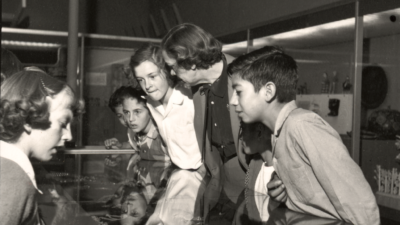 Four children and one adult lean over a glass display case, closely examining the objects inside in a museum or exhibit setting. [gen-ai]