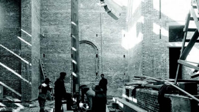 Four people stand inside a partially constructed brick building with beams of sunlight shining through, surrounded by construction materials and equipment. [gen-ai]