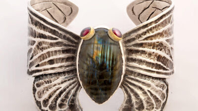 A silver cuff bracelet shaped like butterfly wings, featuring a central iridescent stone and two small red stones as accents. [gen-ai]