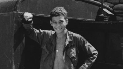 A young man in military uniform stands smiling beside a vehicle with sandbags stacked in the background. [gen-ai]