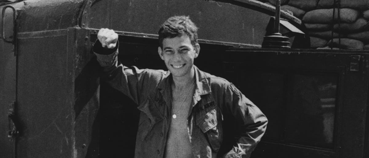 A young man in military uniform stands smiling beside a vehicle with sandbags stacked in the background. [gen-ai]