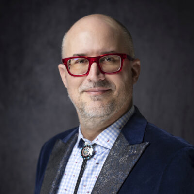A man with glasses, a goatee, and a bald head wears a blue velvet jacket, patterned shirt, and bolo tie against a dark background. [gen-ai]
