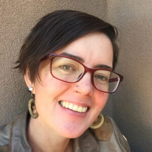 A person with short dark hair, red glasses, and large earrings smiles at the camera while standing against a textured wall. [gen-ai]