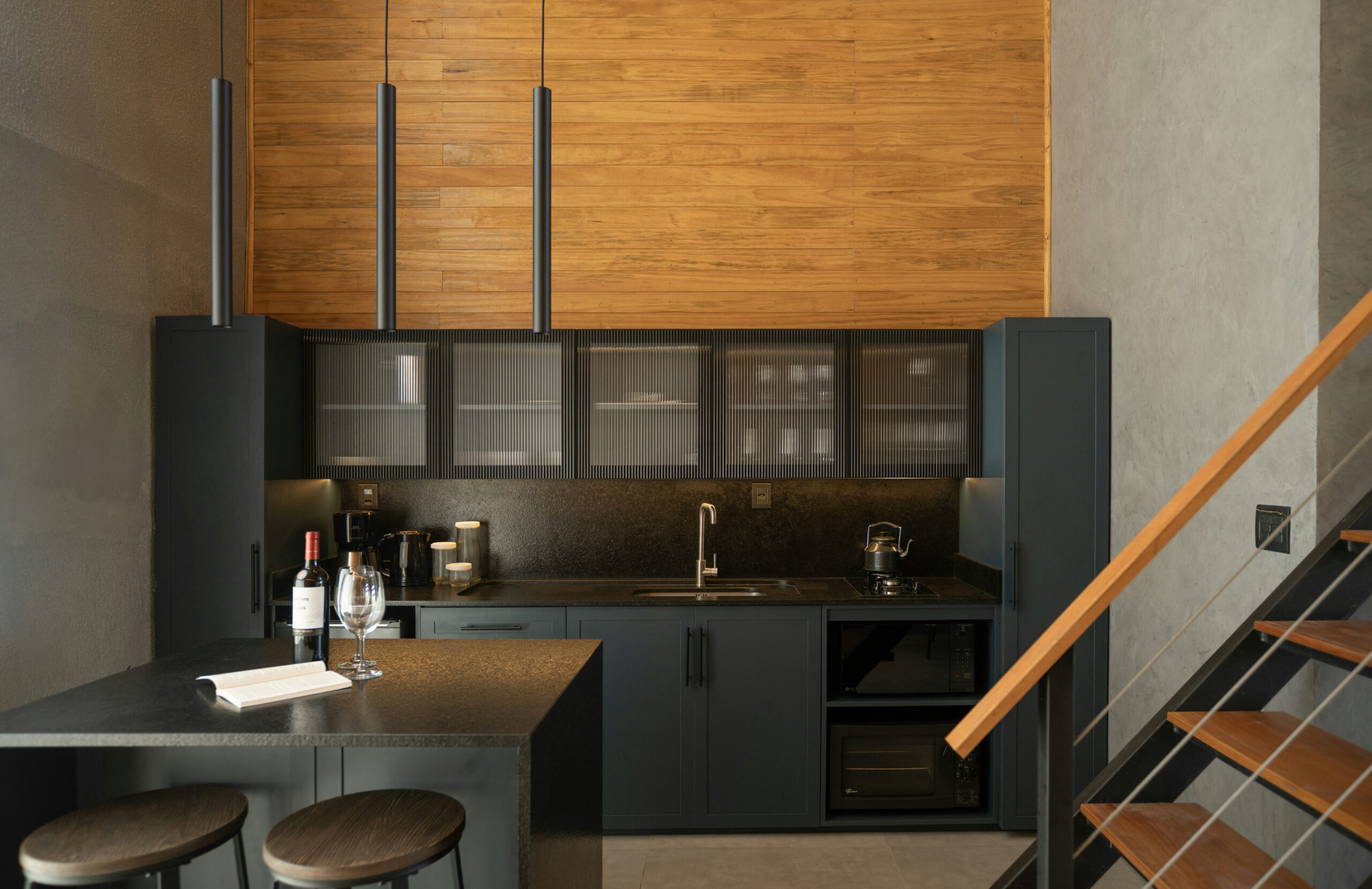 Modern kitchenette with black cabinets, a small bar area with two stools, a bottle of wine with glasses, wooden accents, and a staircase on the right. [gen-ai]