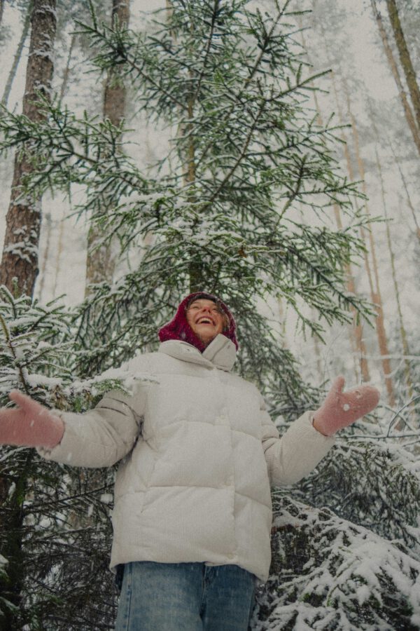 A person wearing a white puffer jacket, red hat, and pink gloves stands in a snowy forest, looking up with arms outstretched. [gen-ai]