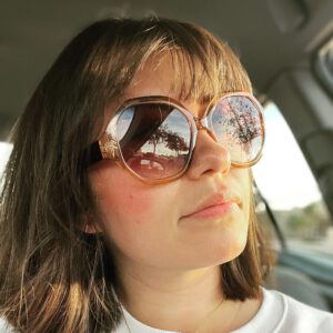 Woman with brown hair and large round sunglasses sitting in a car. Trees and sky are reflected in her sunglasses. She is looking slightly to the side. [gen-ai]