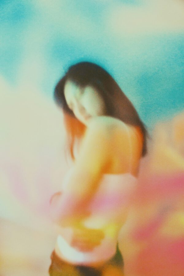 A woman in a white tank top stands against a blurred, colorful background with soft blue, pink, and orange hues. [gen-ai]